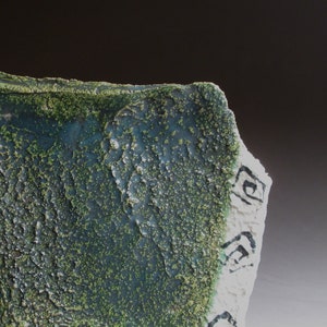 May include: Close-up of a textured ceramic piece with a rough, dark teal surface speckled with green. The edge is white with black spiral designs. The piece has a rugged, organic appearance.