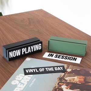 Puede incluir: Porta-carteles negros y verdes con texto blanco. El porta-carteles negro dice "NOW PLAYING" y el porta-carteles verde dice "IN SESSION". Un cartel negro con texto blanco que dice "VINYL OF THE DAY" está encima de un disco de vinilo.