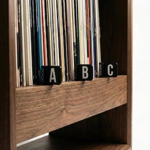 May include: A wooden record shelf filled with vinyl records. Black dividers labelled A, B, and C are placed in front of the records. The shelf is made of dark wood with a visible wood grain.
