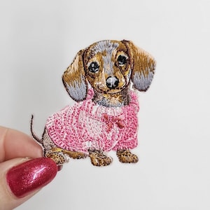 Pode incluir: Patch bordado de um dachshund usando um suéter rosa. O cão tem pelo marrom e cinza com uma expressão doce. O patch é detalhado com costuras intrincadas, perfeito para adicionar a roupas ou acessórios.
