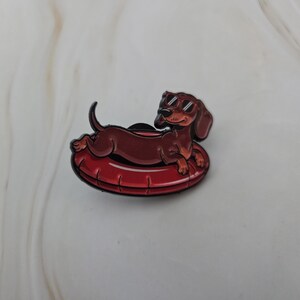 Op de afbeelding: Een cartoon-stijl pin met een bruine teckel met zonnebril, liggend op een rode opblaasbare zwemband. De pin heeft een glanzende afwerking en is een leuk accessoire voor hondenliefhebbers.