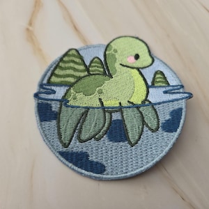 Op de afbeelding: Een ronde geborduurde patch met een lichtgroen wezen met een lange nek en vinnen dat in blauw water zwemt. Het ontwerp bevat donkergroene bergen op de achtergrond, een klein roze wangdetail op het wezen en een donkerblauwe rand.