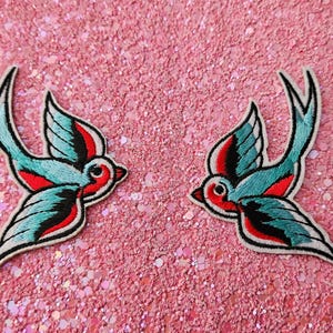 Op de afbeelding: Twee geborduurde zwaluw-patches met een levendig kleurenpalet van lichtblauw, felrood en zwart garen. Elke vogel is afgebeeld in een dynamische vlieghouding met gespreide vleugels, voorzien van gedetailleerd stikwerk en een strakke witte rand.
