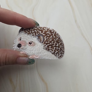 Op de afbeelding: Een geborduurde opstrijkbare patch in de vorm van een egel. Het ontwerp heeft een wit gezicht en buik met een bruine, getextureerde rug die stekels voorstelt. De egel heeft een kleine zwarte neus, een donker oog en lichtroze accenten op de snuit.