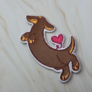 Op de afbeelding: Geborduurde patch met een bruine teckel en een roze hart. De hond staat in een speelse pose, met een witte rand en gele accenten op zijn oren, poten en staart. Ideaal voor het versieren van kleding of accessoires.