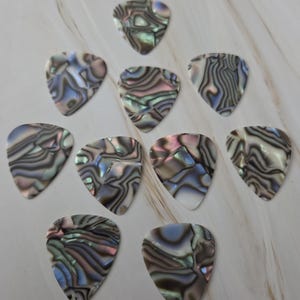 Op de afbeelding: Een verzameling van tien iriserende gitaar plectrums. Elke plectrum heeft een uniek wervelend patroon van kleuren, waaronder blauw, groen en roze, wat een glinsterend effect creëert. De plectrums liggen op een lichtgekleurd oppervlak.