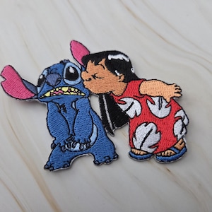 Op de afbeelding: Een geborduurde patch met een blauw buitenaards personage met grote oren en een meisje met zwart haar in een rode jurk met een wit bladpatroon. Het meisje kust het blauwe personage op de wang, alles uitgevoerd in borduurgaren.