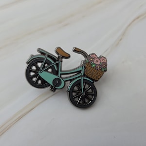 Op de afbeelding: Een turquoise fietsspeld met een bruin zadel en stuur. De fiets heeft zwarte wielen en een mandje gevuld met roze en paarse bloemen. De speld is een decoratief accessoire.