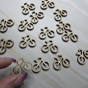 以下が含まれることがあります： 小さなレーザーカットされた木製の自転車の形のコレクション。自転車は薄茶色で、シンプルな二輪のデザインです。画像には、明るい色の表面に散らばっている複数の自転車の切り抜きが写っています。