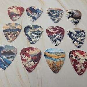 Op de afbeelding: Een verzameling van twaalf gitaar plectrums met Japanse kunst. Elke plectrum toont een ander landschap met golven, bergen en bomen in blauwe, rode en bruine tinten. De plectrums liggen op een lichte ondergrond.