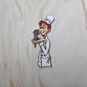 Può includere: Una toppa ricamata raffigurante uno chef dei cartoni animati con capelli castano-rossicci, che indossa un'uniforme bianca e un cappello alto, mentre tiene un piccolo topo grigio sul palmo della mano. Il design è realizzato con fili dettagliati.