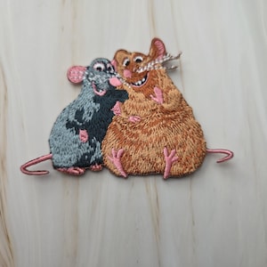Op de afbeelding: Een geborduurde patch met twee cartoonratten die naast elkaar zitten. De rat aan de linkerkant is grijs met roze oren en pootjes, terwijl de grotere rat aan de rechterkant bruin is met een brede grijns, een roze neus en roze pootjes.