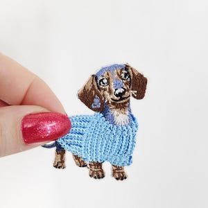 May include: Embroidered dachshund dog patch wearing a light blue sweater. The dog has brown and blue fur with a white muzzle. The patch is held against a white background.