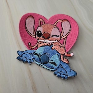 Peut inclure: Un écusson brodé thermocollant représentant un personnage bleu allongé avec un personnage orange clair assis sur son dos, le tout devant une forme de cœur rose uni. Les personnages ont de grandes oreilles, des yeux et des nez foncés.