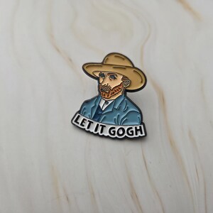 Peut inclure: Une broche en émail métallique représentant une illustration stylisée d'un homme à la barbe brun-roux portant un chapeau beige et une veste bleue. Sous la figure, une bannière blanche avec le texte noir LET IT GOGH en majuscules.