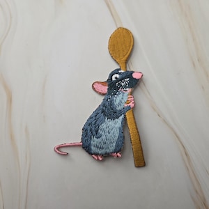 Op de afbeelding: Een geborduurde patch met een donkergrijze rat met roze oren en een roze neus, rechtopstaand en een lange, mosterdgele houten lepel vasthoudend. De rat heeft een lange, dunne staart die naar links uitsteekt en is in zijaanzicht afgebeeld.