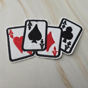 Può includere: Un set di quattro toppe ricamate a forma di carte da gioco, tra cui l'asso di cuori, picche, quadri e fiori. Ogni toppa presenta uno sfondo bianco, un bordo nero e il simbolo corrispondente al seme della carta raffigurata.