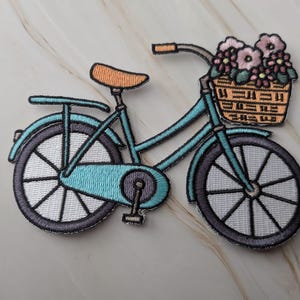 Op de afbeelding: Een geborduurde patch met een blauwgroene fiets met een beige zadel en een mandje aan de voorzijde gevuld met roze en witte bloemen. De fiets heeft donkergrijze wielen met witte spaken, een blauwgroen frame en een beige mandje.