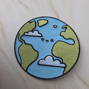 Op de afbeelding: Geborduurde patch met een gestileerd aarde-ontwerp. De patch heeft een blauwe achtergrond die de oceanen voorstelt, met groene continenten en witte wolken. Een zwarte rand omlijnt de cirkelvorm.