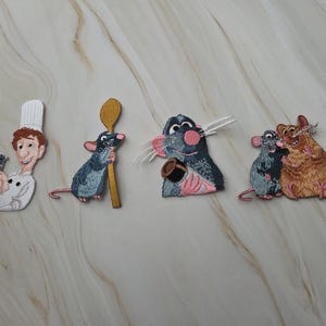 Op de afbeelding: Vier geborduurde patches met geanimeerde figuren: een kok in een wit uniform die een kleine rat vasthoudt, een grijze rat met een gouden lepel, een grijze rat met een kleine bruine pot, en twee ratten, een grijze en een bruine, die samen staan.