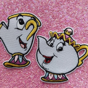Op de afbeelding: Twee geborduurde stoffen patches met stripfiguren in de vorm van een theekopje en een theepot. Het theekopje is wit met gele accenten en een paarse voet. De theepot is wit met een geel handvat, een roze deksel en een paarse voet.