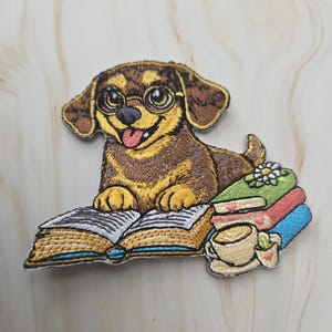 Op de afbeelding: Geborduurde patch met een bruine en gele hond met een bril, zittend op een open boek. Een stapel boeken en een kopje thee staan naast de hond. De hond steekt zijn tong uit.