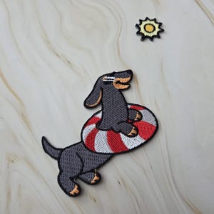Op de afbeelding: Geborduurde patch met een teckel met een zonnebril en een rood-wit gestreepte zwemband. Er is ook een kleine geel-zwarte zon-patch aanwezig. De patches liggen op een lichtgekleurd oppervlak.