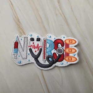 Op de afbeelding: Een geborduurde patch met het woord NURSE in gestileerde letters. De N is een injectiespuit, de U een stethoscoop, de R een thermometer, de S een reflexhamer en de E bestaat uit drie oranje pillen. De achtergrond is wit met blauwe hartjes.