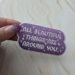 Op de afbeelding: Een paarse geborduurde patch met de woorden "ALL BEAUTIFUL THINGS ARE AROUND YOU" in het wit. De patch heeft een afgeronde rechthoekige vorm met kleine ster- en stipaccenten.