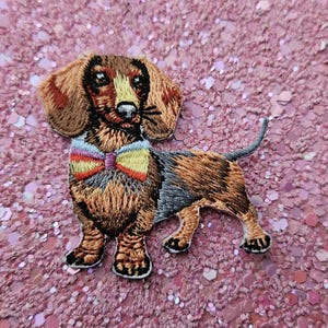 Könnte beinhalten: Gestickter Aufnäher eines Dackelhundes mit braunem, schwarzem und hellbraunem Fell, der eine bunte Fliege trägt. Der Aufnäher befindet sich auf einem rosa Glitzerhintergrund. Der Hund steht.