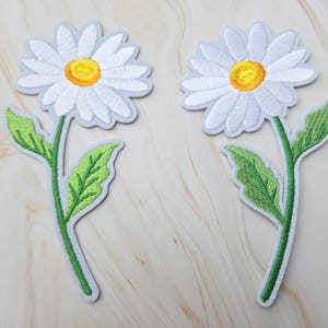 Op de afbeelding: Twee geborduurde madeliefje-patches met witte bloemblaadjes, een felgeel hart en een groene steel met twee bladeren. Elke patch heeft een strakke witte rand en is ontworpen om als decoratief bloemenaccent op stoffen te worden aangebracht.