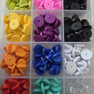 Op de afbeelding: Een doorzichtige plastic organizer gevuld met kleurrijke pin backs. De pin backs zijn in verschillende kleuren, waaronder groen, roze, zwart, geel, paars, oranje, blauw, wit, rood en teal. Elke pin back heeft een ronde bovenkant en een pin.