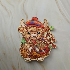 Può includere: Una toppa ricamata raffigurante una soffice mucca Highland marrone che indossa un berretto a quadri rossi e blu. La mucca suona una cornamusa con una sacca a quadri rossi e verdi, arricchita da piccoli dettagli a forma di trifoglio verde.