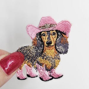 Może przedstawiać: Haftowana naszywka przedstawiająca jamnika w różowym kapeluszu kowbojskim i różowych kowbojskich butach. Pies ma brązowe, czarne i żółte futro. Naszywka jest na białym tle.