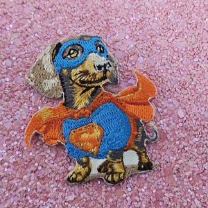 Könnte beinhalten: Gestickter Aufnäher eines Dackelhundes als Superheld. Der Hund trägt eine blaue Maske und ein Brustemblem mit einem orangefarbenen Umhang. Auf rosa Glitzerhintergrund.