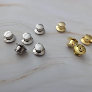 Op de afbeelding: Een verzameling zilver- en goudkleurige pin backs, verspreid op een lichtgekleurd, gemarmerd oppervlak. De pin backs zijn klein, rond en hebben een pen- en sluitingontwerp, geschikt voor het vastzetten van pins of broches.