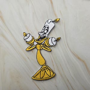 Op de afbeelding: Een geborduurde patch met een geel en wit antropomorf kandelaarfiguur met een lachend gezicht, twee armen die brandende kaarsen vasthouden en een brede voet, uitgevoerd in levendig geel, wit, zwart en oranje garen op een stoffen ondergrond.