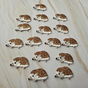 Op de afbeelding: Een verzameling van veertien kleine, geborduurde egel-patches gerangschikt in een driehoekige formatie. Elke patch heeft een bruine, getextureerde achterkant die stekels voorstelt, een crèmekleurig gezicht en een klein zwart neusje en oogje.