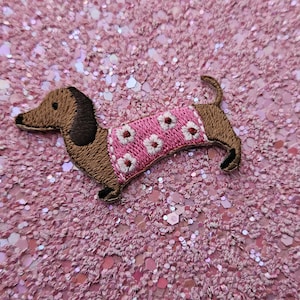 May include: Embroidered dachshund patch in brown and black, wearing a pink sweater with white flower details. The patch is set against a sparkly pink background.