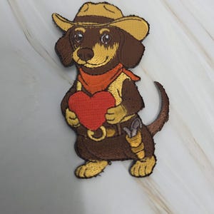 May include: Embroidered patch featuring a dachshund wearing a cowboy hat and a red bandana. The dog is holding a red heart and has a gun in a holster. The patch is brown, yellow, and red.