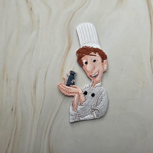 Op de afbeelding: Een geborduurde patch met een kok met bruin haar, een witte koksmuts en een wit koksbuis. De kok houdt een kleine donkergrijze rat in de open handpalm. Het ontwerp maakt gebruik van gedetailleerd stikwerk voor de gelaatstrekken en kleding.