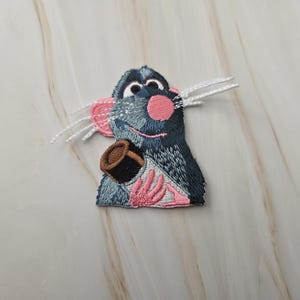 Op de afbeelding: Een geborduurde patch met een donkerblauwe rat met een roze neus, roze oren en roze handjes. De rat heeft grote ogen en lange witte draadsnorharen en houdt een klein bruin en wit voorwerp met beide handen tegen de borst aan.