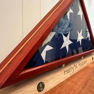 Puede incluir: Un expositor de madera para banderas con frente de cristal, que muestra una bandera estadounidense plegada. La caja es de madera de color marrón rojizo con una base de color claro. El nombre "Harry N. Russos" y las fechas están grabados en la base.
