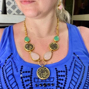 May include: Gold-toned necklace with coin pendants, teardrop-shaped pink stones, and green stones. Matching earrings with pink and green stones. The jewelry is worn over a blue top.