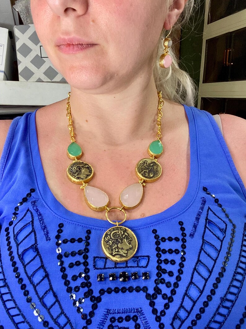 May include: Gold-toned necklace with coin pendants, teardrop-shaped pink stones, and green stones. Matching earrings with pink and green stones. The jewelry is worn over a blue top.