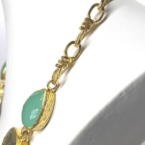 May include: Gold-toned necklace with a chain design and teardrop-shaped, light green gemstones. The necklace is displayed on a white mannequin bust. The gemstones are set in textured gold bezels.