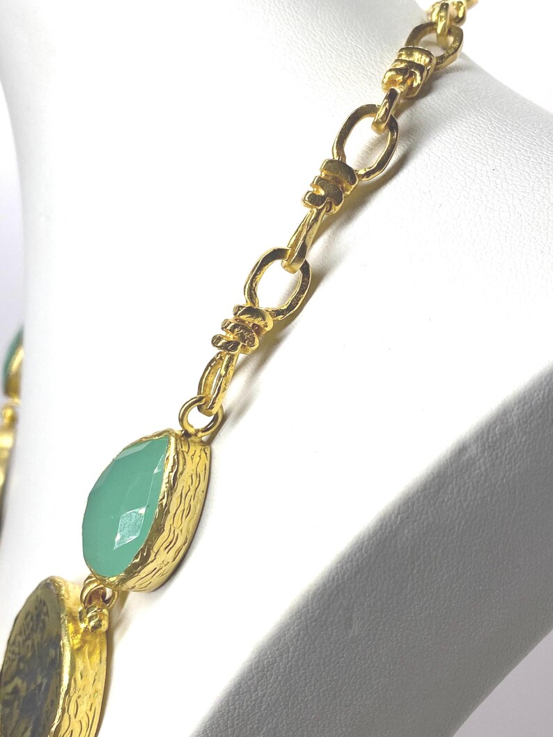 May include: Gold-toned necklace with a chain design and teardrop-shaped, light green gemstones. The necklace is displayed on a white mannequin bust. The gemstones are set in textured gold bezels.