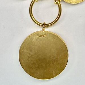 May include: A gold-toned necklace with a large circular pendant. The pendant has a textured surface and the word "TURKEY" is stamped on the back. The necklace features a large circular ring and smaller circular accents.
