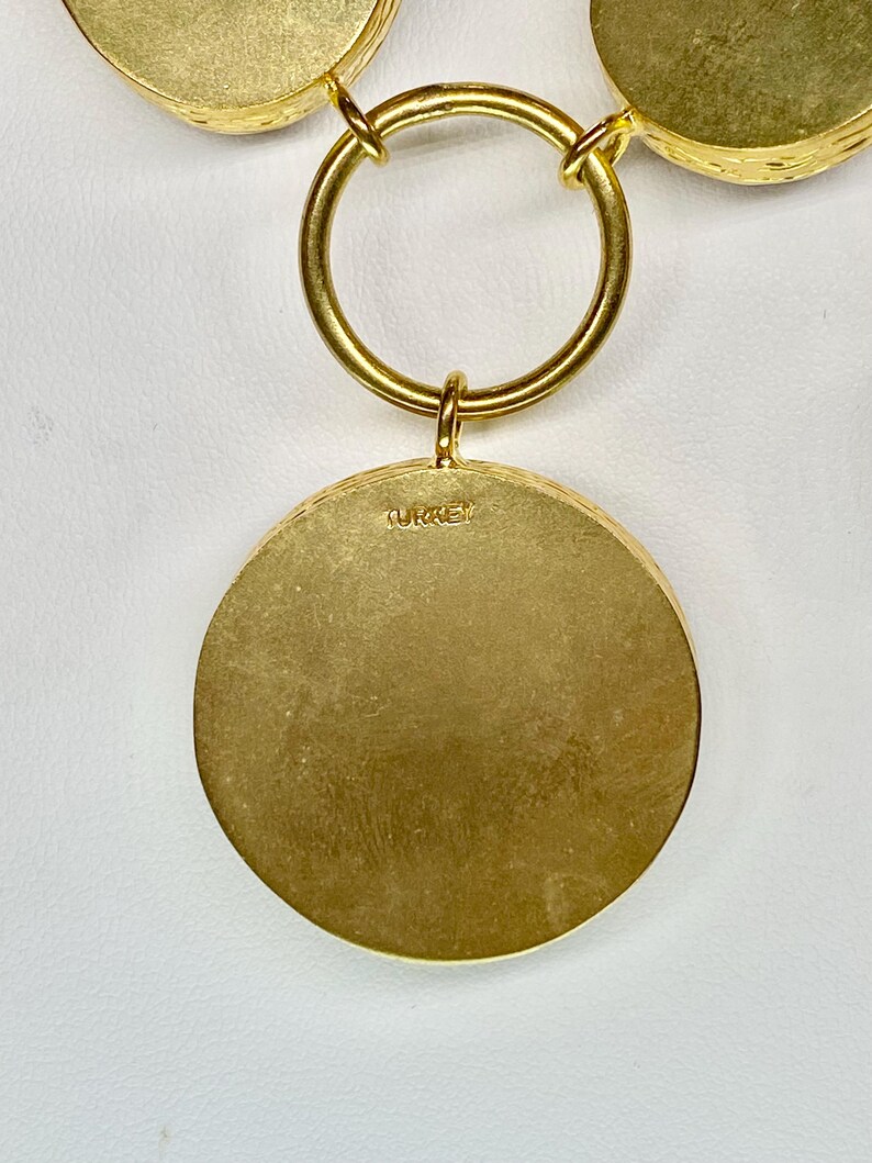 May include: A gold-toned necklace with a large circular pendant. The pendant has a textured surface and the word "TURKEY" is stamped on the back. The necklace features a large circular ring and smaller circular accents.