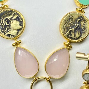 May include: Gold-toned necklace featuring coin-shaped pendants with embossed figures, paired with teardrop-shaped pink gemstones. The necklace includes a matching earring with a small, round, light-colored stone.